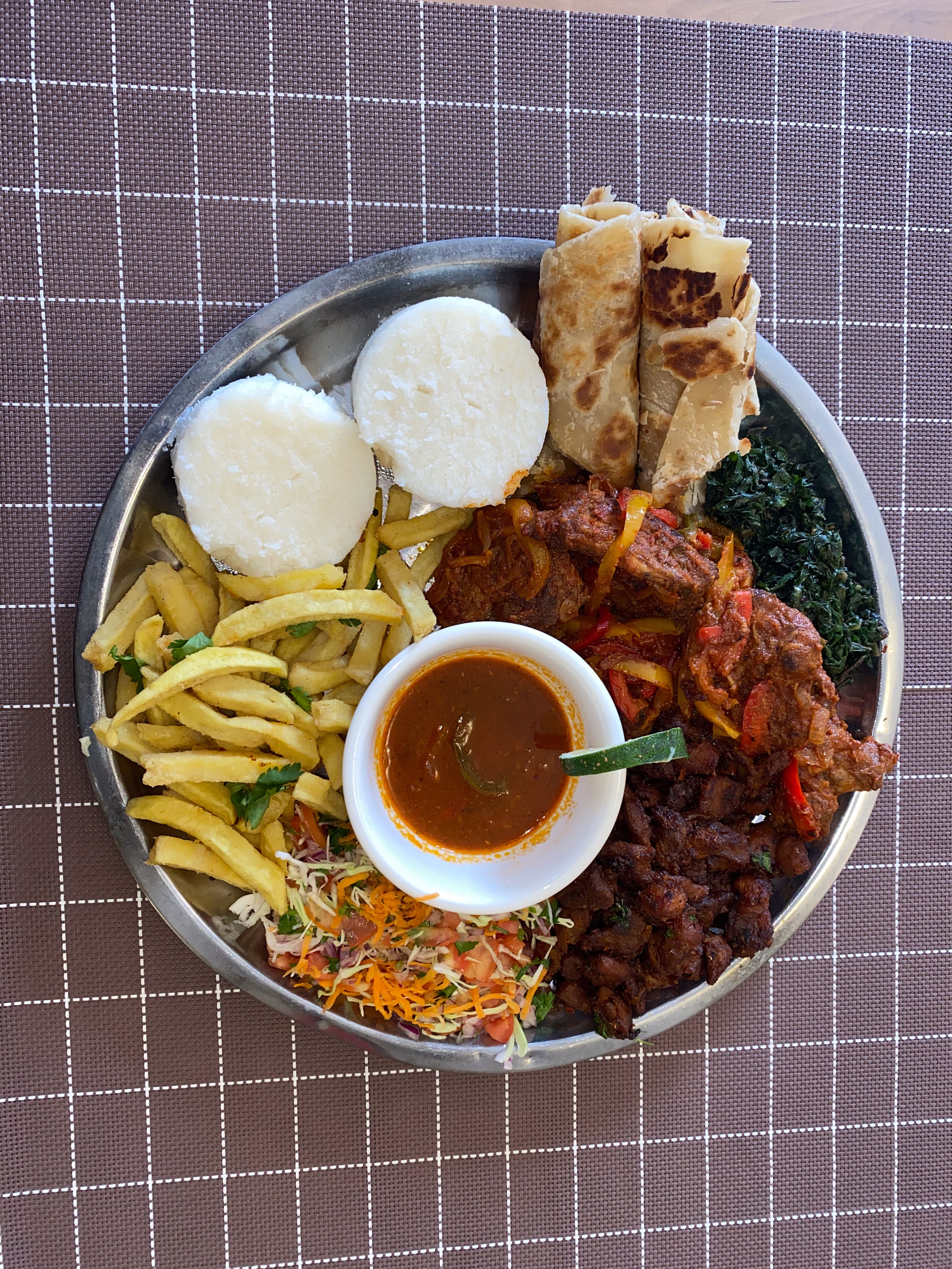 A sharing platter from above — ugali, chapati rolls, fries, greens and two meat stews around a bowl of sauce