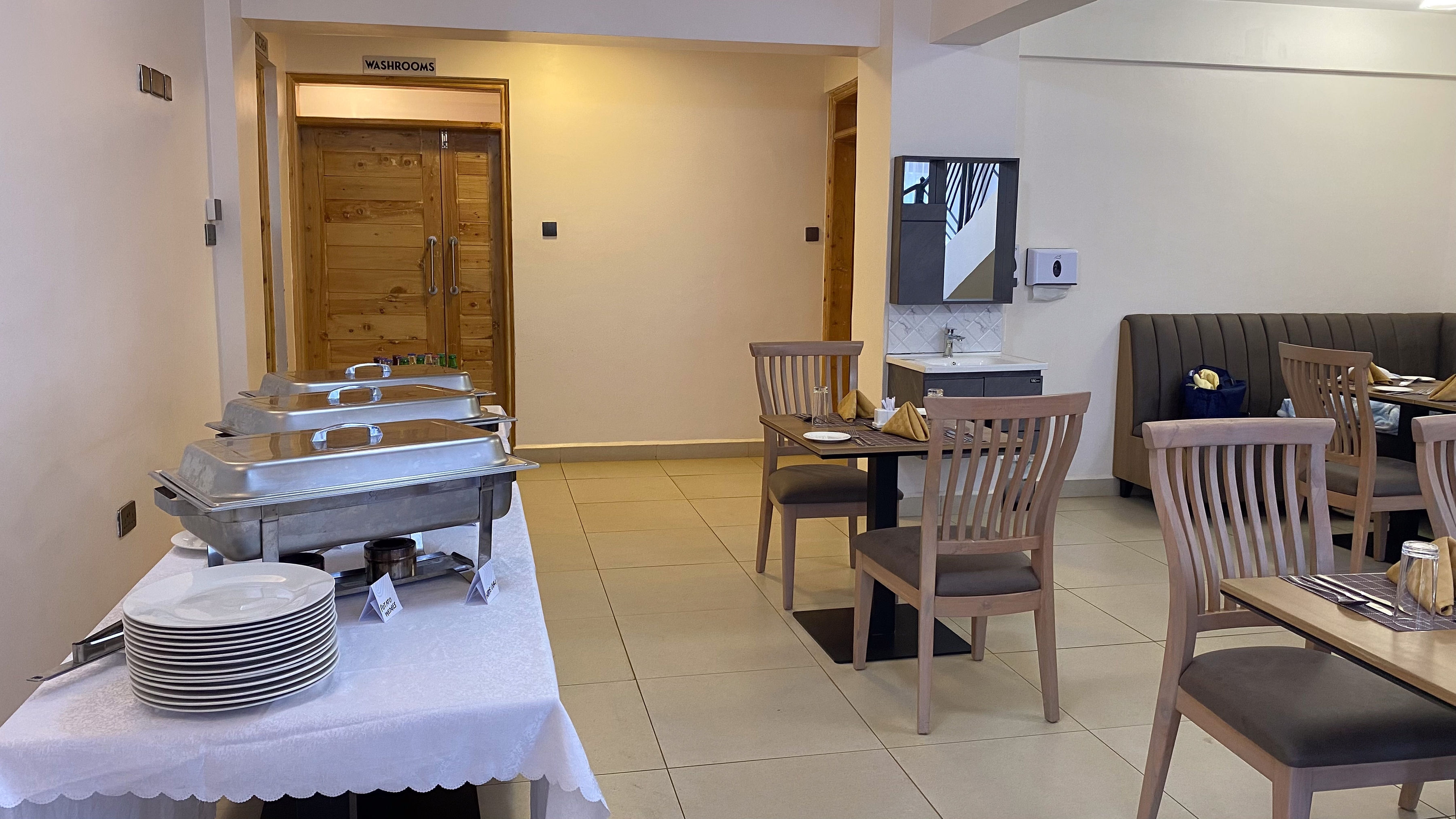 Wooden dining chairs and set tables beside a buffet station with covered serving dishes
