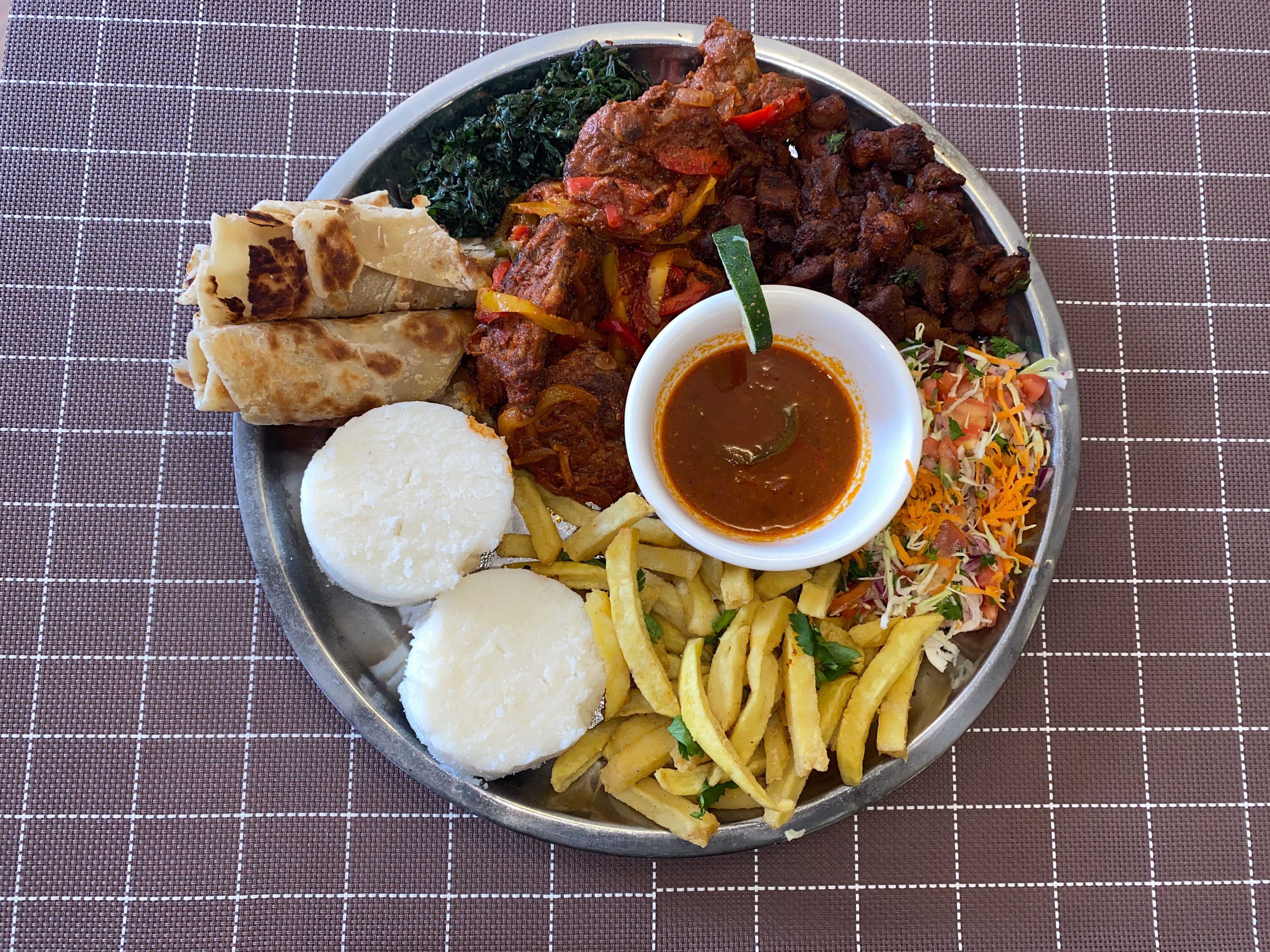 A round sharing platter — fried beef bites, stewed meat with peppers, greens, ugali rounds, chapati rolls, fries and a bowl of sauce