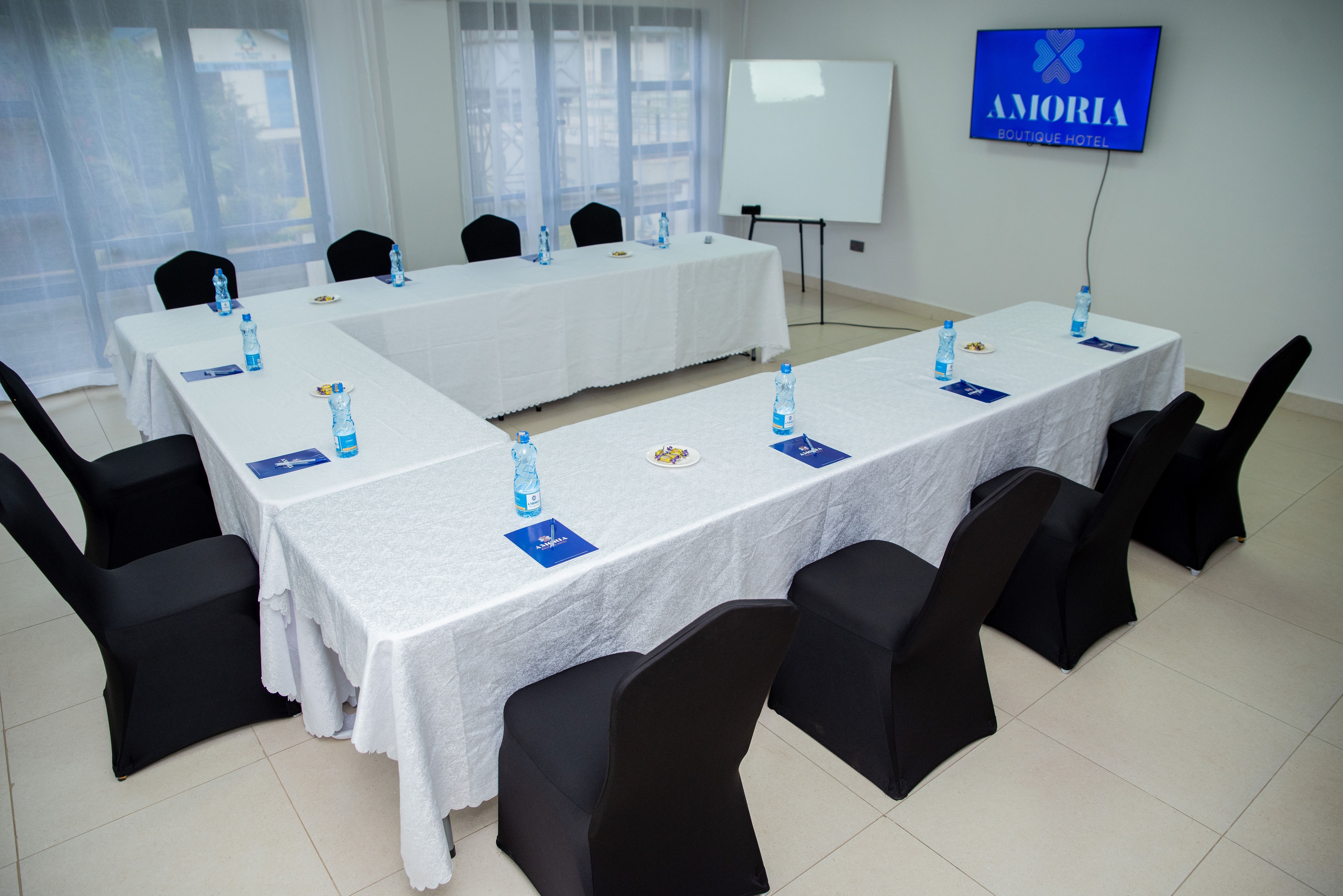 Conference tables set with bottled water and notepads beside sheer-curtained windows and black covered chairs