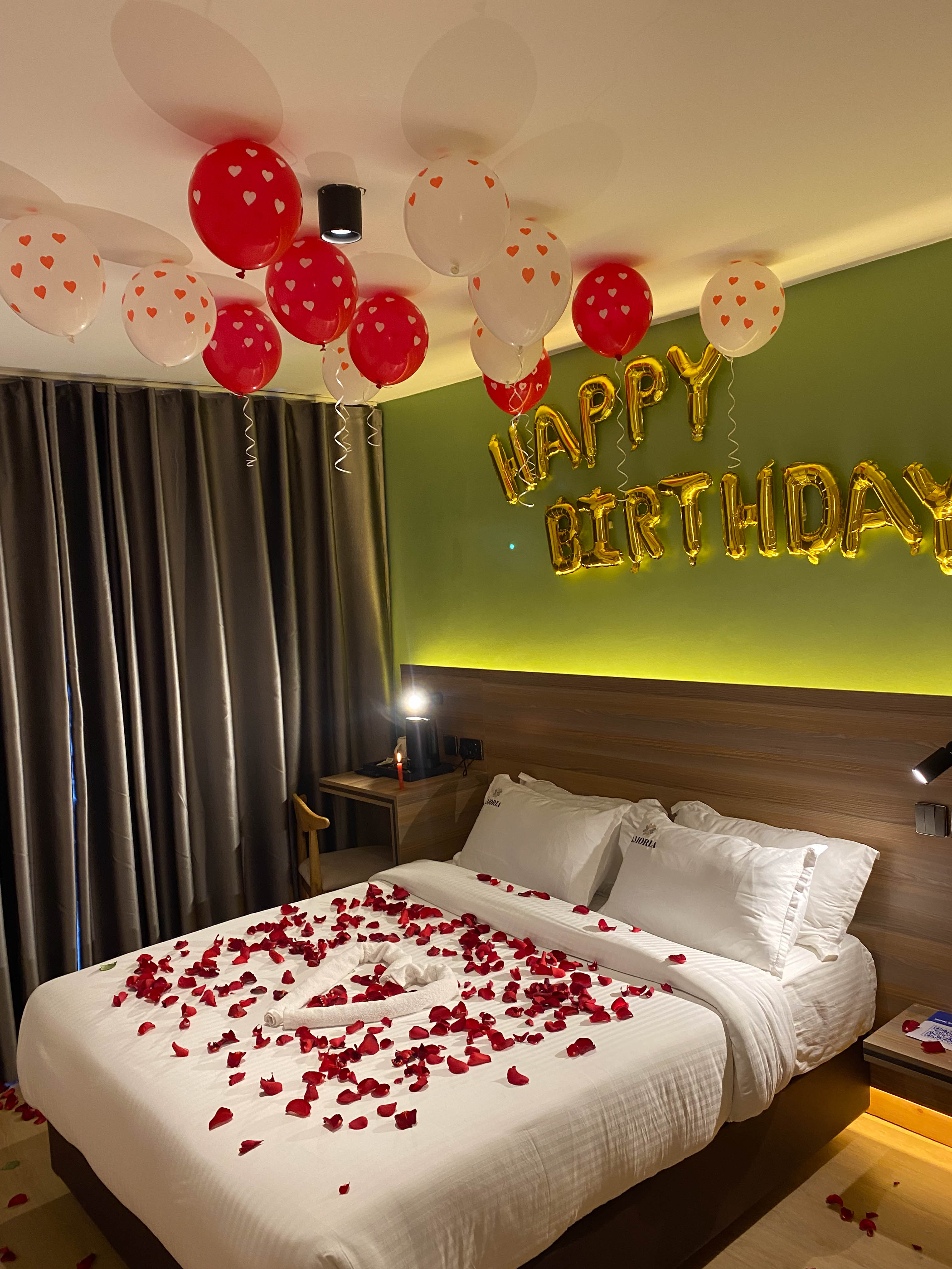 Deluxe room staged for a birthday — gold foil letters and heart balloons over a rose-petal-strewn bed