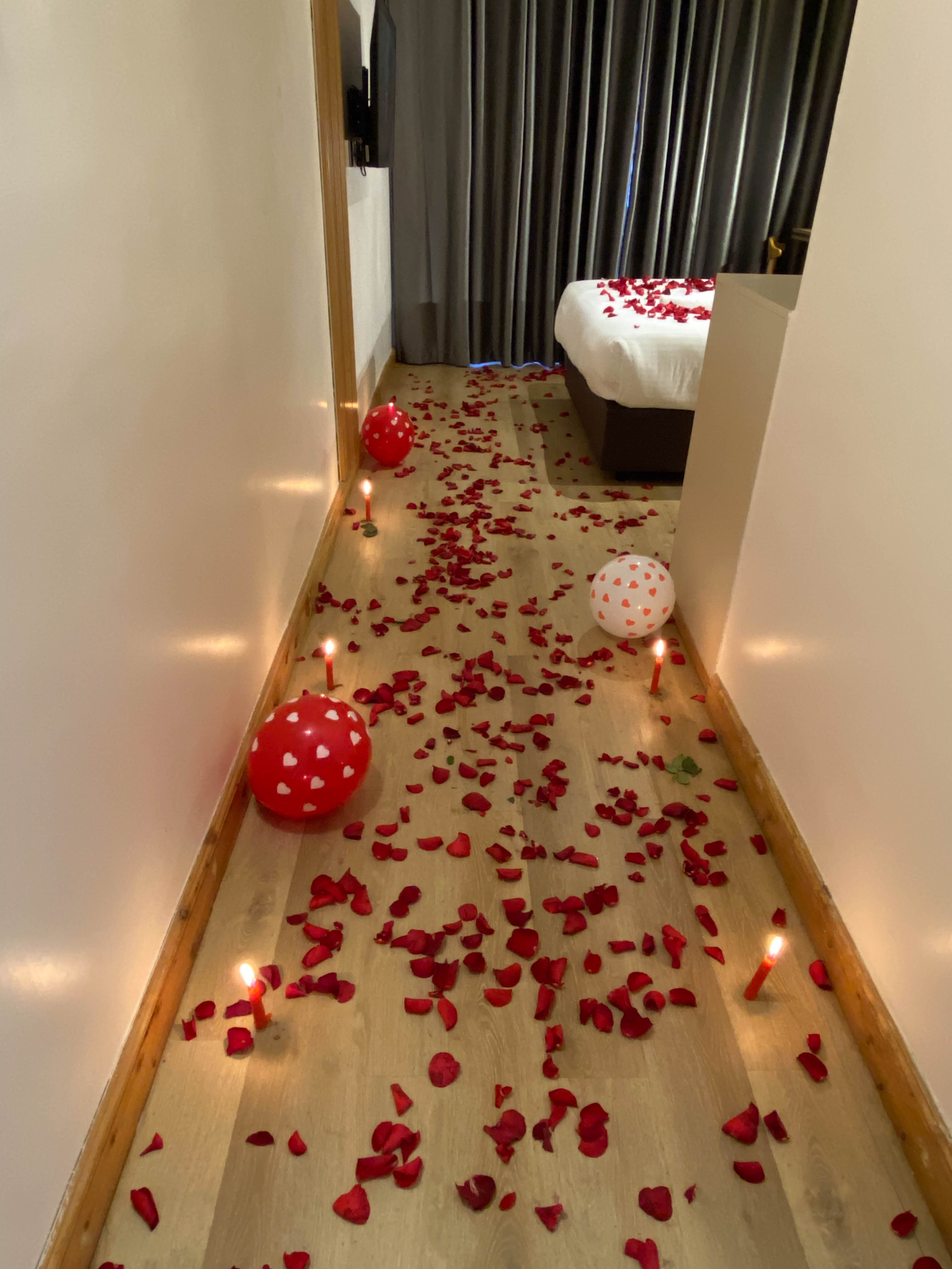Trail of rose petals, candles and heart balloons leading into a room with a petal-covered bed