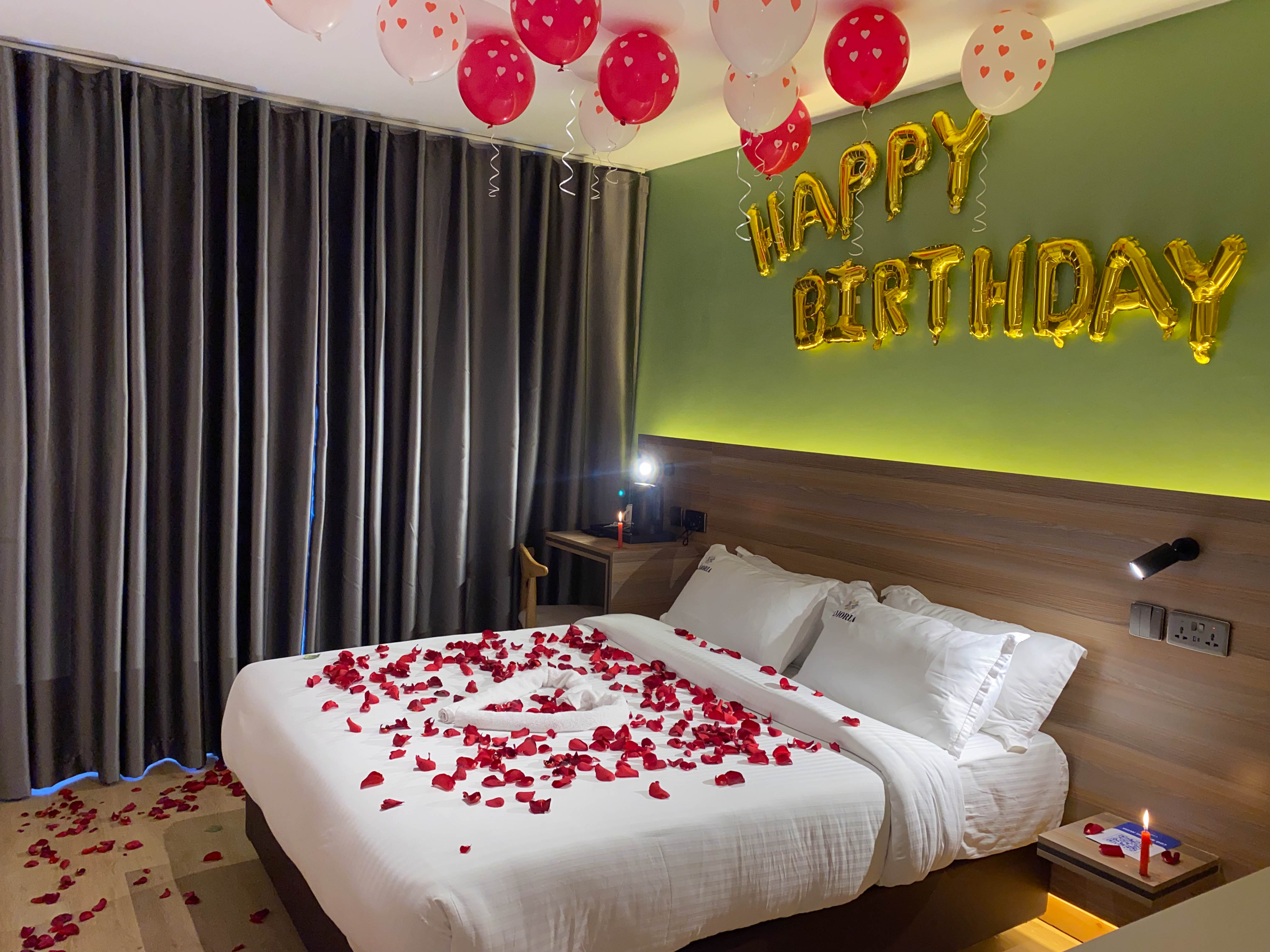 Room staged for a birthday — heart balloons and gold foil letters over a bed scattered with rose petals