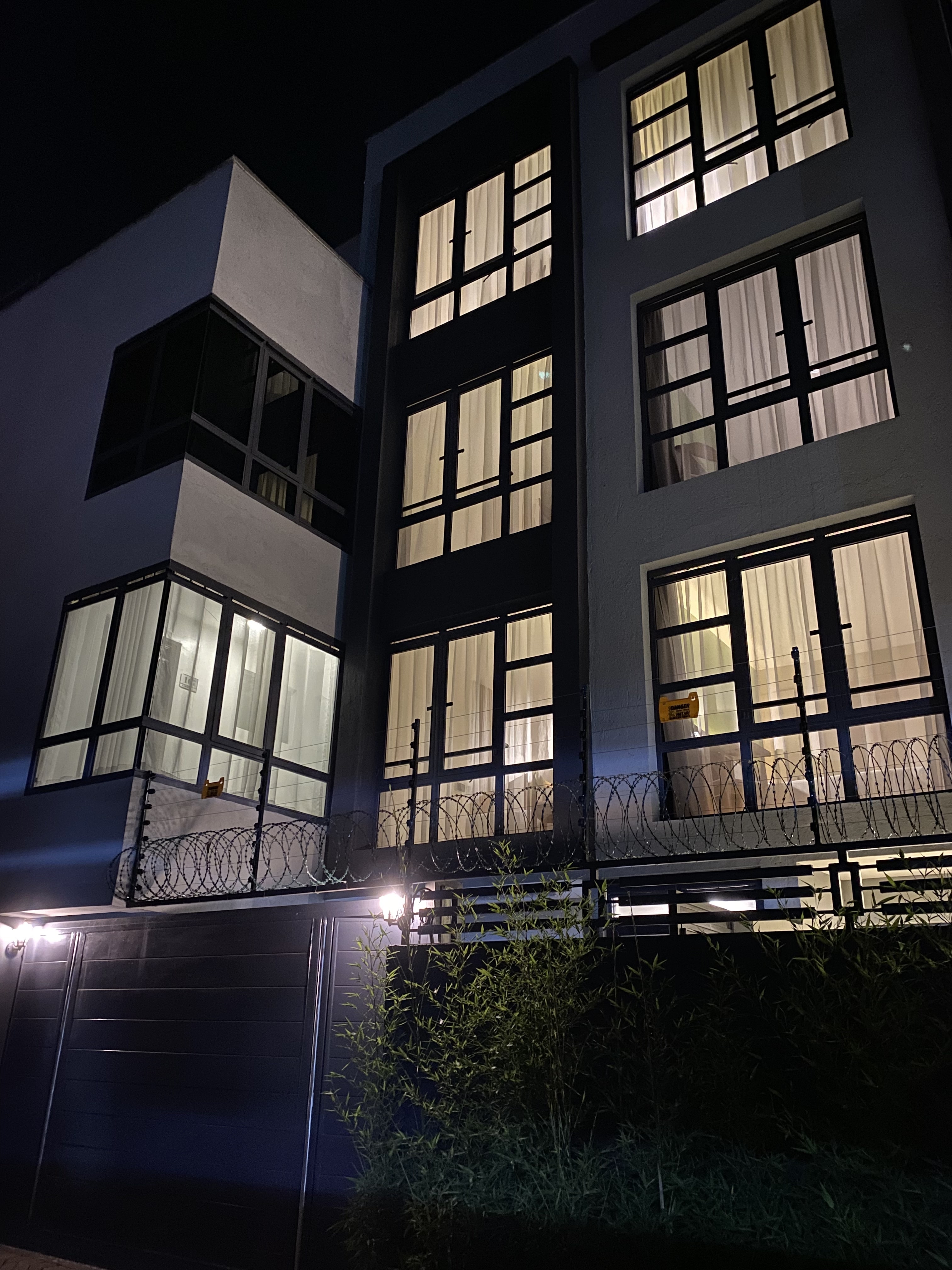 The hotel facade after dark, rows of windows glowing warm behind sheer curtains