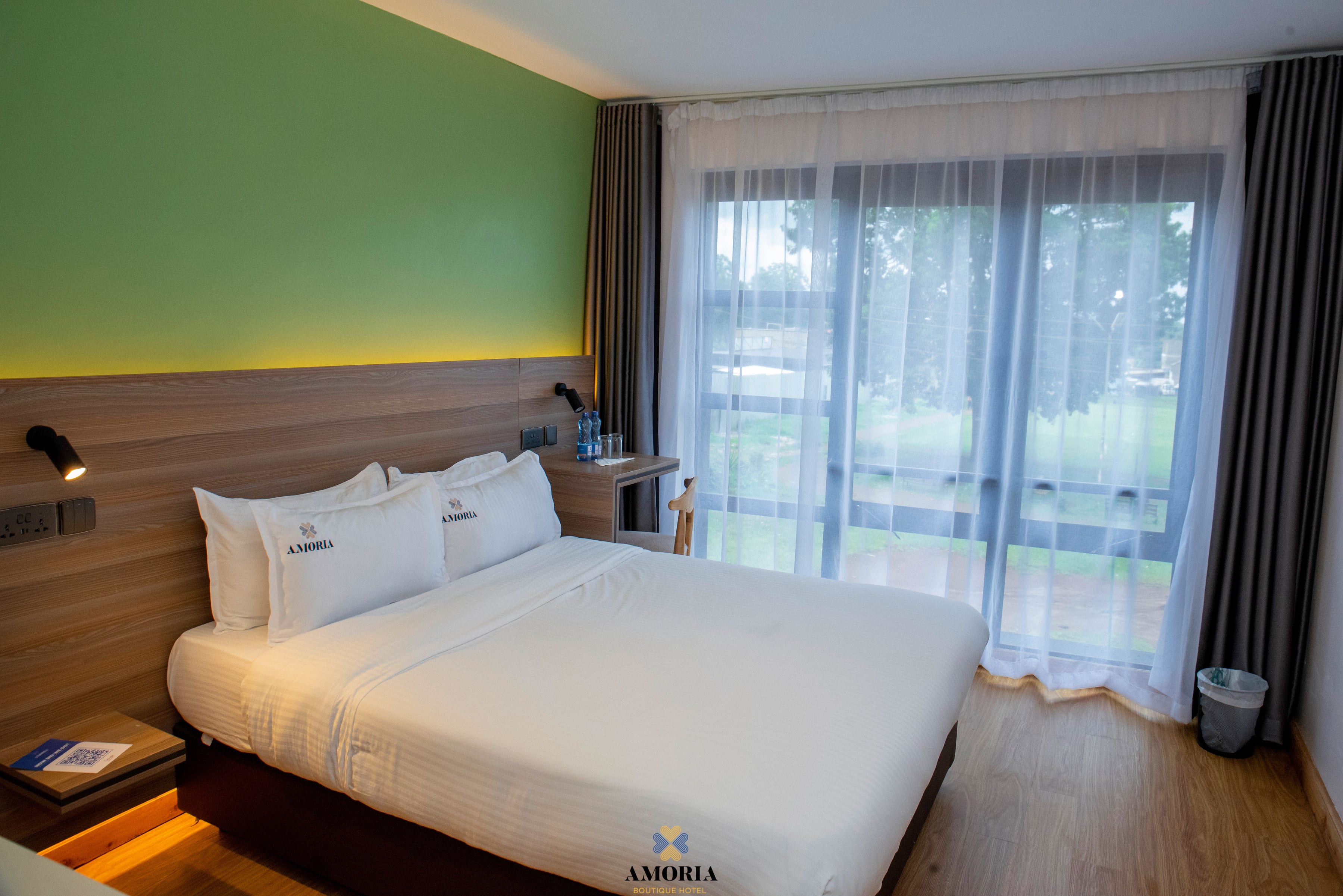 Double bed with Amoria-embroidered pillows beside floor-to-ceiling sheers, the lawns green beyond the glass