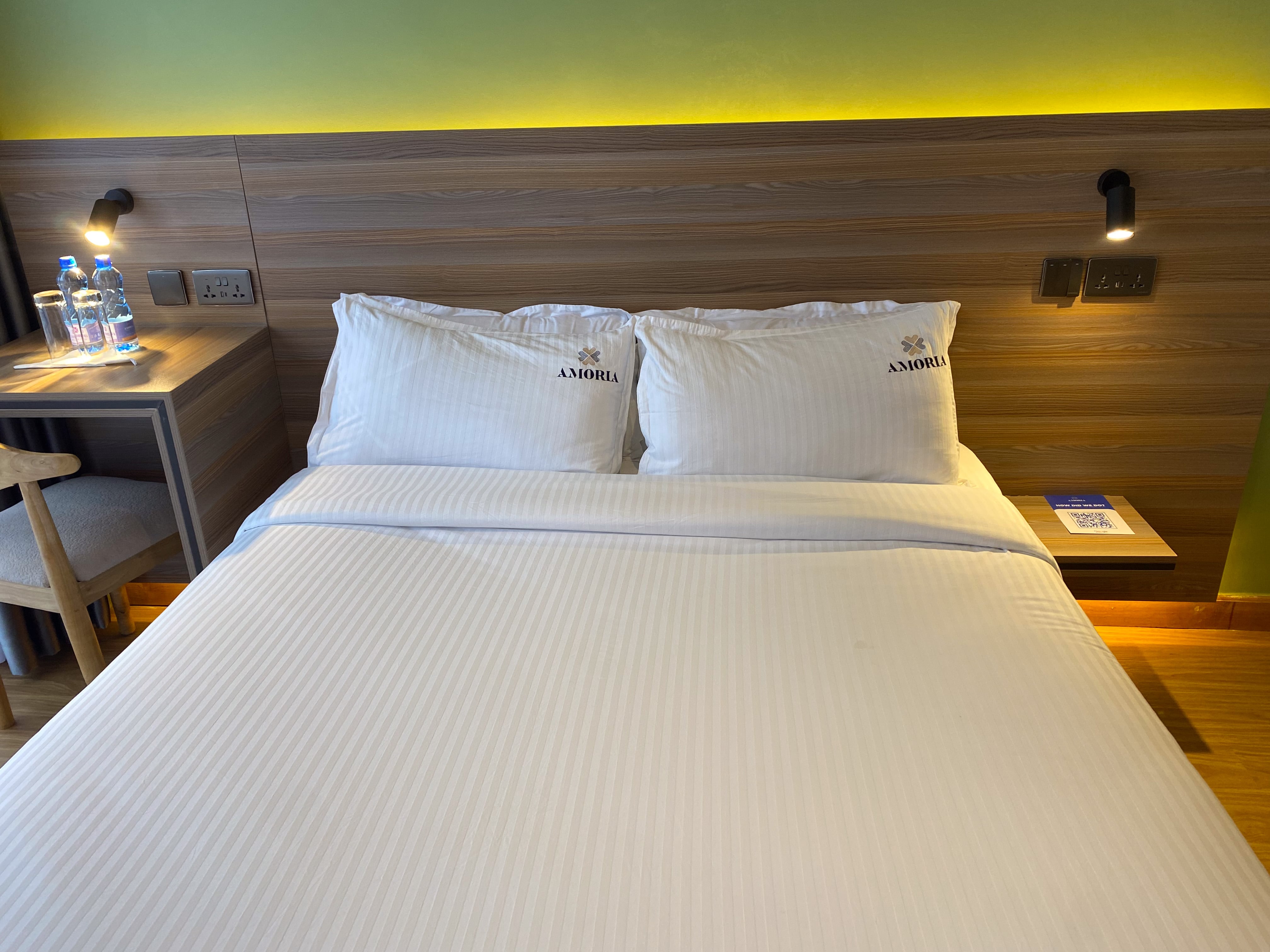 The Standard room's double bed head-on — Amoria-embroidered pillows, backlit headboard and water bottles on the desk