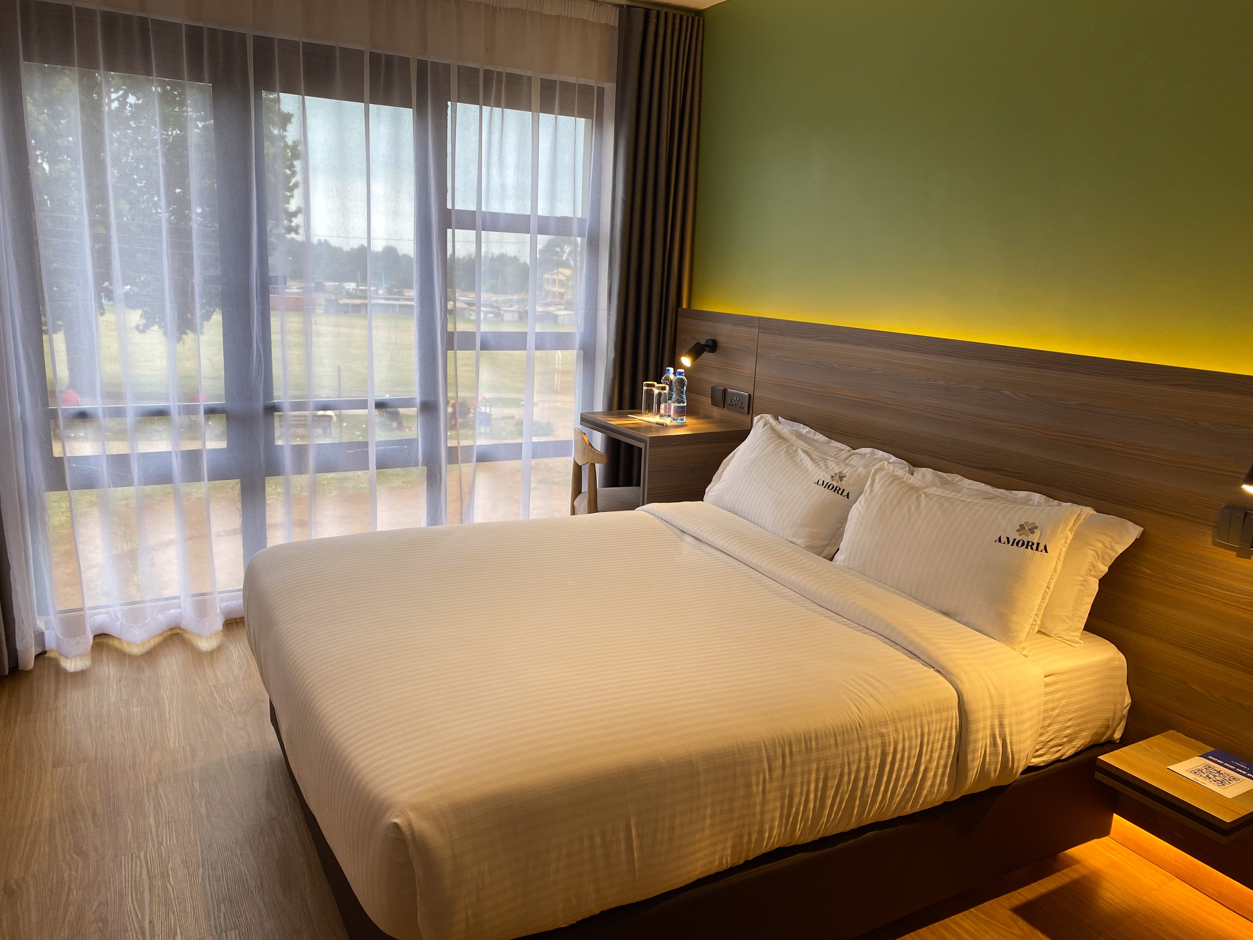 Standard room with a made double bed beside a floor-to-ceiling sheer-curtained window, the green feature wall glowing above the headboard