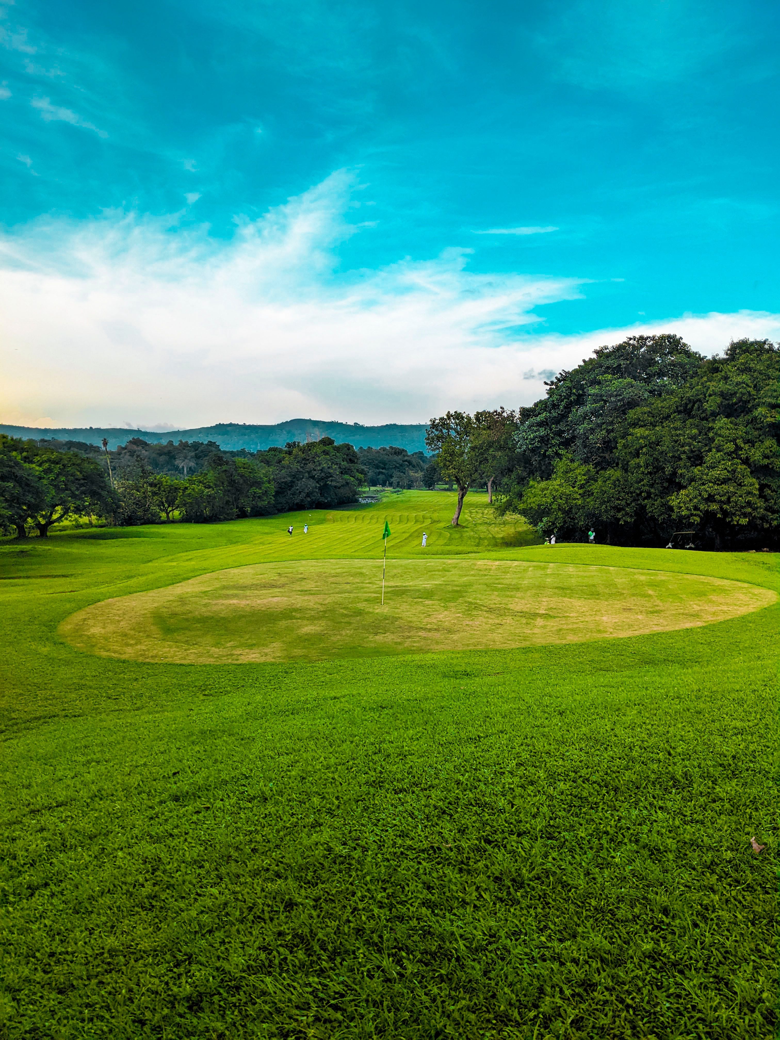 A golf green with its flag under a wide blue highland sky, players and a cart out along the tree-lined fairway